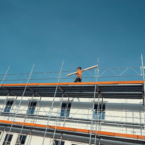 En byggarbetare som bär på planka gåendes på byggnadsställning